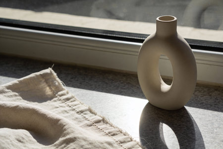 A modern, matte beige ceramic vase with a unique oval opening is placed on a windowsill next to a...