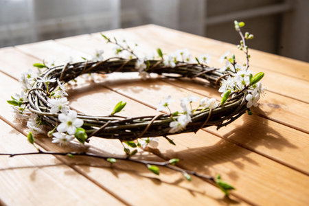 A rustic wreath crafted from natural twigs, decorated with small white flowers and bud leaves, resting on a wooden surfaceの写真素材
