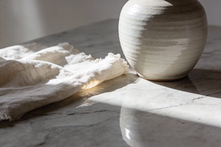 A close-up view captures the textured folds of a natural linen cloth alongside a rustic ceramic vase, illuminated by soft, dappled sunlight on a cool marble countertopの写真素材
