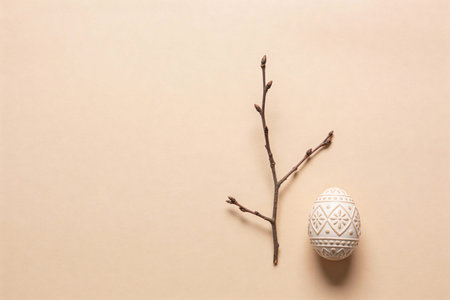 A single decorative Easter egg with intricate patterns sits next to a bare twig on a plain,...