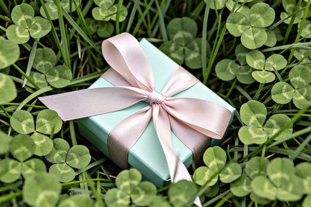 A small, elegant gift box tied with a delicate pink ribbon sits surrounded by vibrant green clover...