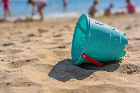 A bright turquoise plastic bucket rests on the sandy shore, its red handle visible, while blurred...
