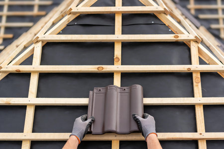 Close-up of a roofer's gloved hands holding a brown ceramic roof tile against a wooden frame and black waterproofing membraneの写真素材
