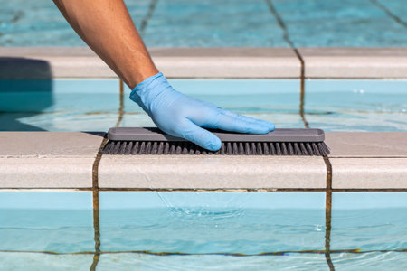 A person wearing blue nitrile gloves uses a stiff brush to scrub the tiled edge of a swimming pool, indicating routine maintenance and cleaningの写真素材