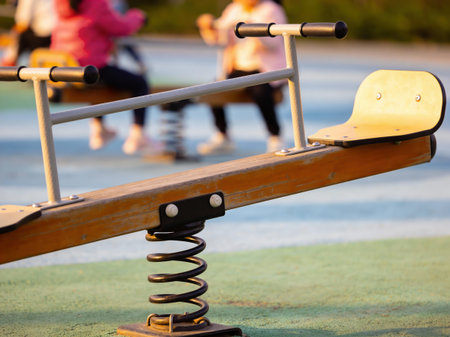A close-up view of a wooden seesaw with a spring mechanism, while blurred figures of children enjoy playtime in the background on a sunny dayの写真素材