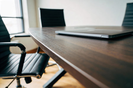 A sleek laptop rests on a polished dark wood conference table surrounded by executive chairs, suggesting a professional meeting spaceの写真素材