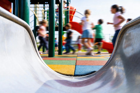 A low-angle view of a metal slide with blurred children running and playing in the background on a bright summer dayの写真素材