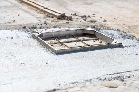 A square metal manhole frame sits open on a rough surface with gravel and wooden planks visible in the background, suggesting ongoing work
