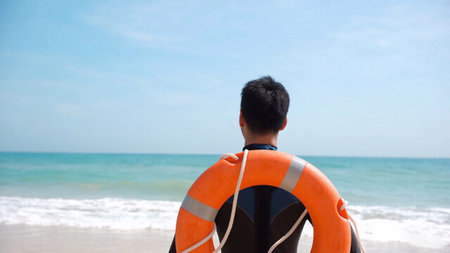 Man in a rescue uniform, standing near the shoreline with the sea behind him. For blogs on emergency preparedness, public safety, working as a lifeguardの写真素材