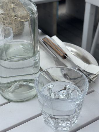 Glass of water with ice cubes on white wooden table next to a glass bottle and cutlery, enhancing...