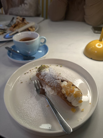 Plate with a cannoli dessert filled with cream and garnished with powdered sugar, next to a cup of...