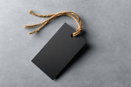 A blank black tag lies on a smooth gray surface, ideal for labeling items or for creative projects. The tag features a twine string loop, enhancing its rustic appearance.の素材