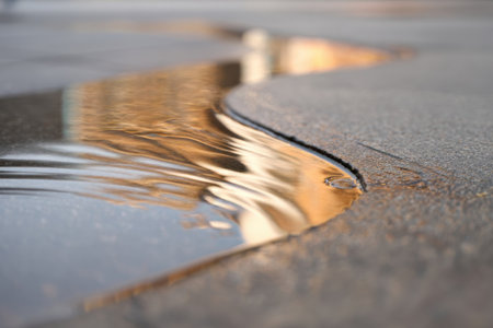 A small puddle on a textured surface reflects warm hues of the sky during golden hour. The sunlight creates a shimmering effect on the waters surface.の素材