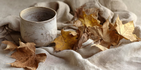 A ceramic cup sits on a soft fabric background, surrounded by dried autumn leaves in shades of brown and yellow, evoking a cozy and serene feeling of the fall season.の素材