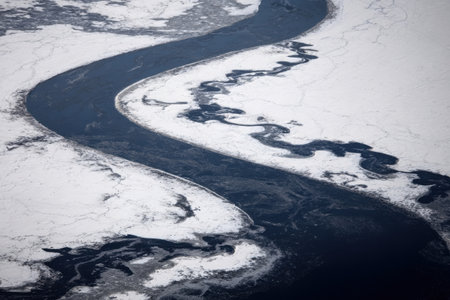 A narrow river snakes through a winter landscape covered in ice and snow. The contrast of dark water against white ice creates a striking visual in the cold season.の素材