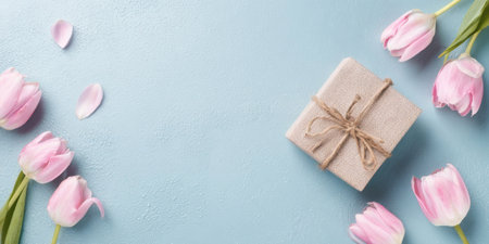 A lovely gift box tied with twine sits on a soft blue surface, accompanied by fresh pink tulips and scattered flower petals creating a cheerful display.の素材