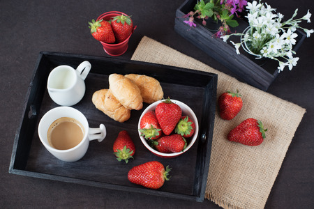 Coffee, mini French pastries and strawberries on wooden tray over black table. White and purple flowers in a decorative wooden crate. Black backgroundの写真素材