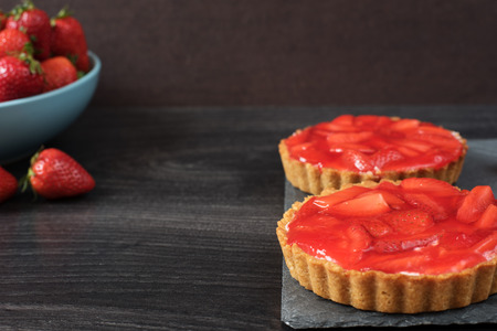 Chocolate tart, tartalette with white chocolate and mascarpone cream, fresh strawberries on top. Large blue bowl full of strawberries. Black wooden background.の写真素材