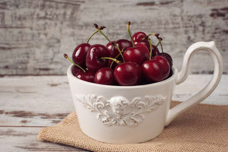 A large cup of coffee in front angel, white bowl full with fresh cherries, fruits. Light rustic background, shabby chic, vintage tintingの写真素材