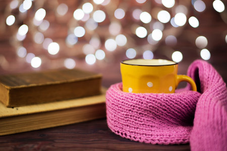 Hot tea, hot chocolate, coffee in yellow cup, wrapped with a pink knitted scarf.  Old books. Blurred...