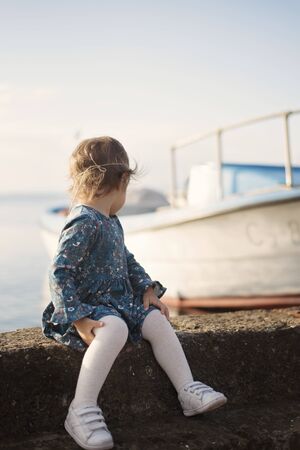 A little girl with a blue dress on the quay looking out to the sea. Boat in the background. Filter, tintingの写真素材