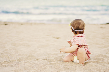 Cute baby girl with a dress on polka dots sitting on the sand on seashore and looks out to the seaの写真素材