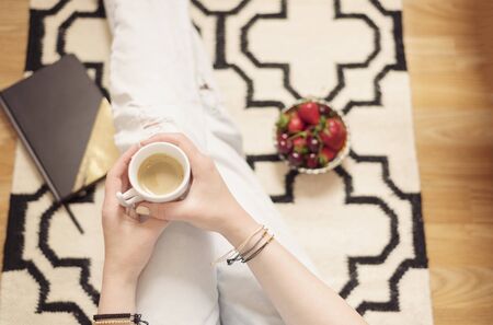 A young woman wearing distressed jeans sitting on wood floor on a rug carpet at home and holding a cup of coffee in hands. Around a golden notebook and a bowl with strawberriesの写真素材