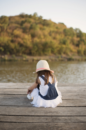 Little girl sitting alone on a wooden bridge by the riverの写真素材