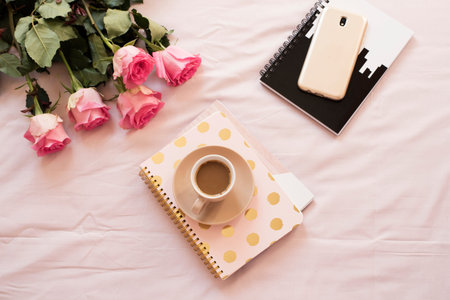 Coffee in bed on pink sheets. Roses, notebooks, gold smartphone around. Freelance fashion home...