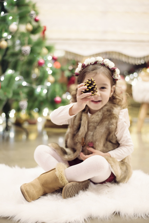 Cute little girl celebrating Christmas at home. She sits on a chair and holds a spruce cone in her hands. Positive emotions. Christmas tree in background. Happy childhood.の写真素材
