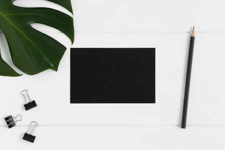 Top view of a black card mockup with workspace accessories and a monstera leaf on a white table.の写真素材