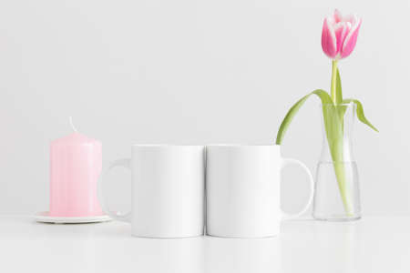 Two mugs mockup with pink tulips in a vase and candle on a white table.の写真素材