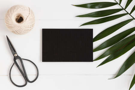 Top view of a black card mockup with workspace accessories and a palm leaf on a white table.の写真素材