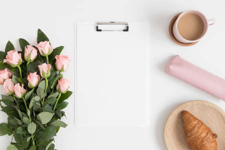 Top view of a white clipboard mockup with a bouquet of pink roses, coffee and a croissant on a white table.の写真素材