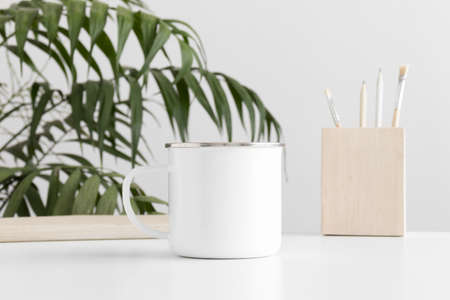 Enamel mug mockup with workspace accessories on a white table and a palm plant.の写真素材