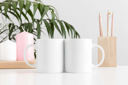 Two mugs mockup with workspace accessories and candles on a white table and a palm plant.の写真素材
