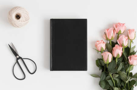 Top view of a black book mockup with a bouquet of pink roses, scissors and twine on a white table.の写真素材