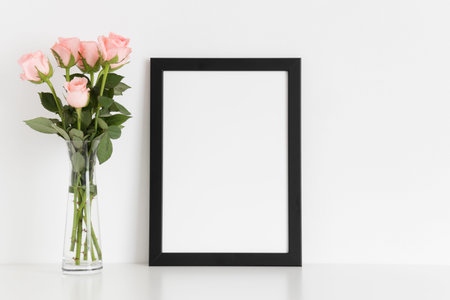 Black frame mockup with a bouquet of pink roses in a glass vase on a white table. Portrait...