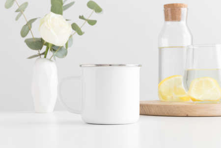 Enamel mug mockup with flowers in a vase, glass and a bottle on a white table.の写真素材