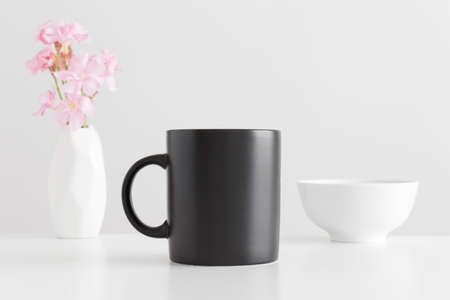 Black mug mockup with a bouquet of a pink oleander in a vase and a bowl on a white table.の写真素材