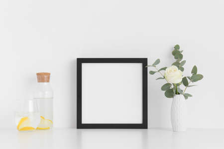 Black square frame mockup with a rose and eucalyptus in a vase, glass and a bottle on a white table.の写真素材