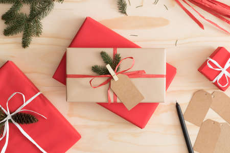 Red and kraft christmas gifts surrounded with pine cones, leaves and twine on a wooden table.