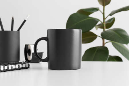 Black mug mockup with workspace accessories on a white table and a ficus plant.の写真素材