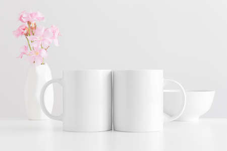 Two white mugs mockup with a bouquet of a pink oleander in a vase and a bowl on a white table.の写真素材