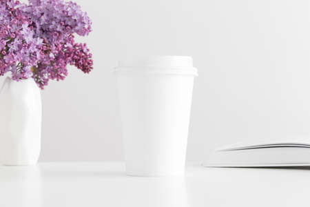 Coffee paper cup mockup with a book and a bouquet of lilac in a vase on a white table.の写真素材