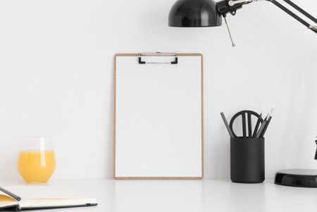 Wooden clipboard mockup wtih a notebook, lamp and workspace accessories on a white table.