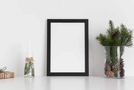 Black frame mockup with a bouquet of fir, candle and a gift on a white table. Christmas decoration.の写真素材
