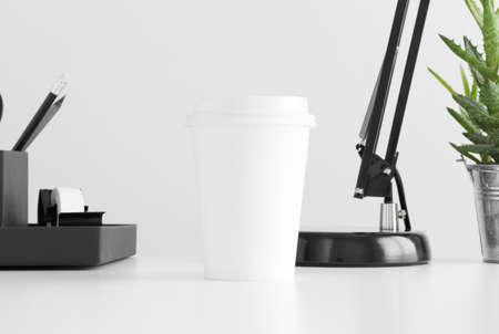 Coffee paper cup mockup with a lamp, workspace accessories and a aloe vera plant in a pot on a white table.の写真素材