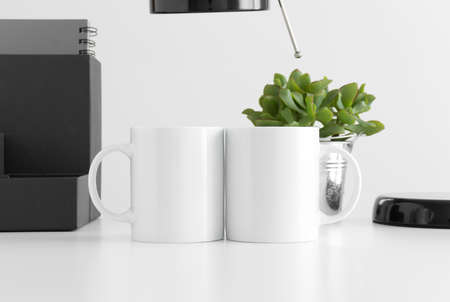 Two white mugs mockup with a lamp, succulent plant and workspace accessories on a white table.の写真素材