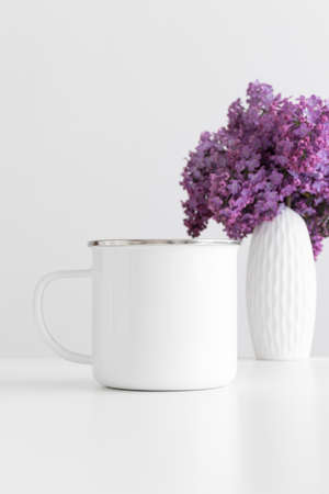 Enamel mug mockup with a bouquet of lilac in a vase on a white table.の写真素材
